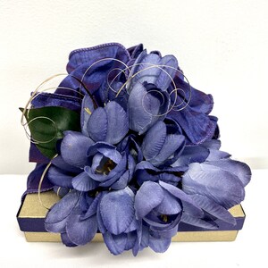 May include: A purple gift box with a bouquet of purple silk flowers on top. The flowers are arranged in a cluster and have a delicate, feminine look.