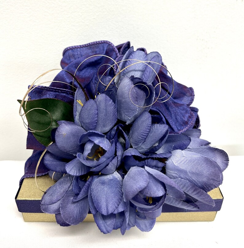 May include: A purple gift box with a bouquet of purple silk flowers on top. The flowers are arranged in a cluster and have a delicate, feminine look.