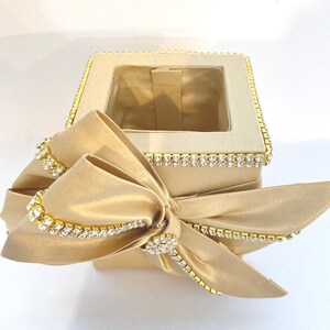 May include: A square-shaped tissue box cover with a gold satin finish and a large bow. The box is decorated with a row of clear rhinestones around the top edge.