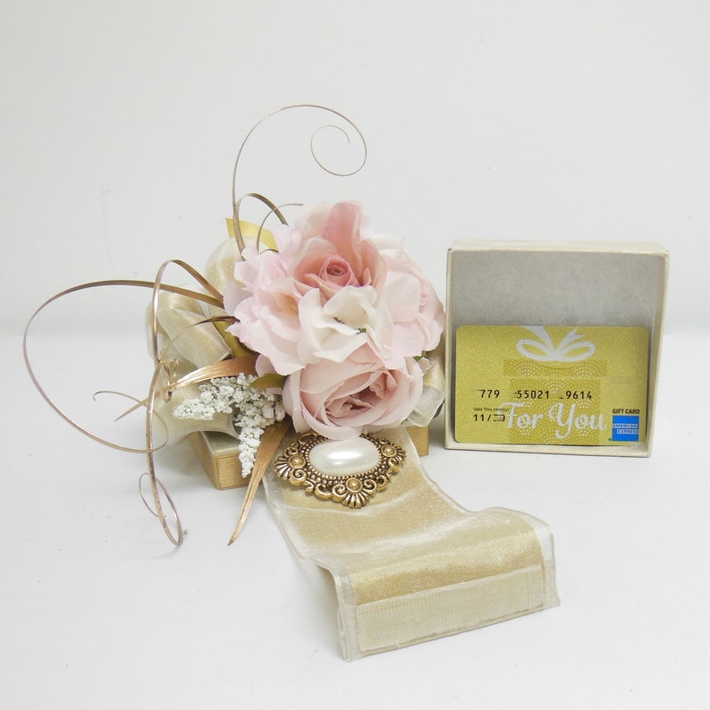 K&ouml;nnte beinhalten: Eine Geschenkbox mit einem rosafarbenen Blumenarrangement und einem goldenen Band. Die Box enth&auml;lt eine Geschenkkarte mit dem Text "F&uuml;r Dich" und einer Geschenkkarten-Nummer.