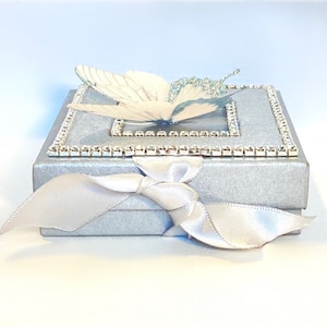 May include: A silver gift box with a white butterfly design and a white satin ribbon. The box is decorated with a row of rhinestones around the top edge.