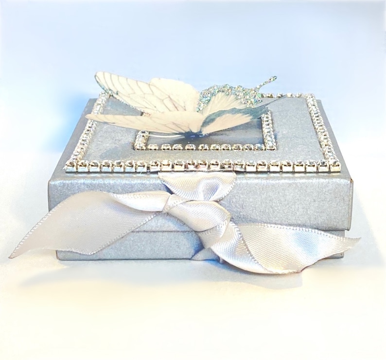 May include: A silver gift box with a white butterfly design and a white satin ribbon. The box is decorated with a row of rhinestones around the top edge.