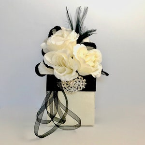 May include: A white gift box with a black ribbon and a bouquet of white roses with a black feather and rhinestone brooch.