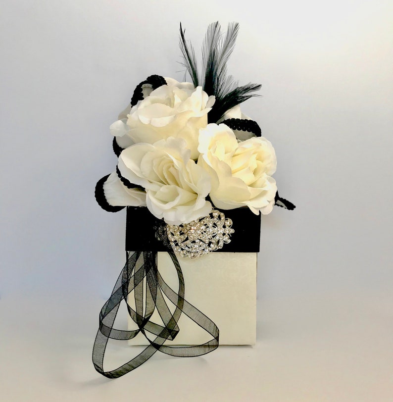 May include: A white gift box with a black ribbon and a bouquet of white roses with a black feather and rhinestone brooch.
