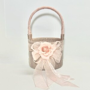 May include: A small, decorative basket with a handle, covered in sparkling rhinestones. A large, light pink fabric rose and matching ribbon bow adorn the front. The handle and bow are a soft pink color. This basket is ideal for weddings or special events.