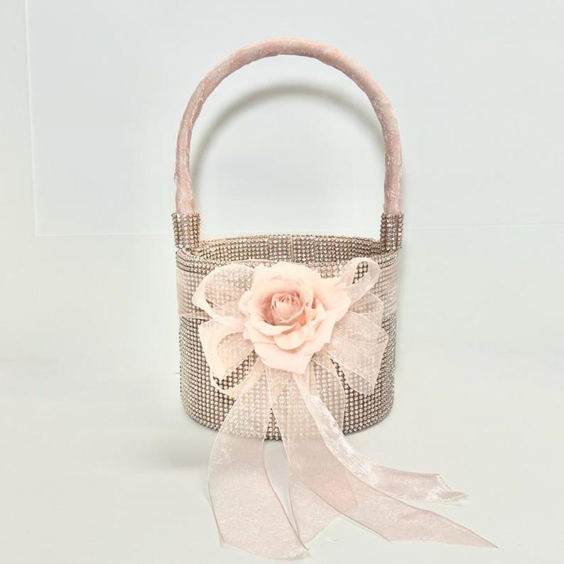 May include: A small, decorative basket with a handle, covered in sparkling rhinestones. A large, light pink fabric rose and matching ribbon bow adorn the front. The handle and bow are a soft pink color. This basket is ideal for weddings or special events.