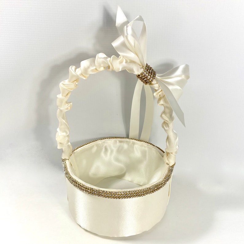May include: A white satin flower girl basket with a white satin bow and a rhinestone embellishment. The basket has a round base and a handle.