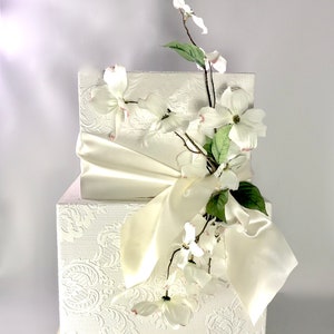 May include: A white two-tiered box with a white satin ribbon and a sprig of white flowers.