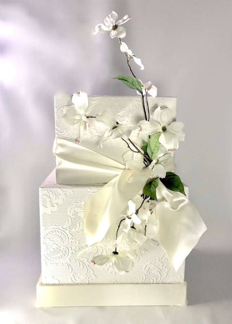 May include: A white two-tiered box with a white satin ribbon and a sprig of white flowers.