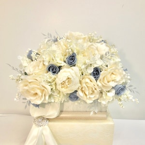 Pu&ograve; includere: Un arrangiamento floreale bianco e blu in una scatola bianca. L'arrangiamento comprende rose bianche, peonie bianche e ortensie blu.