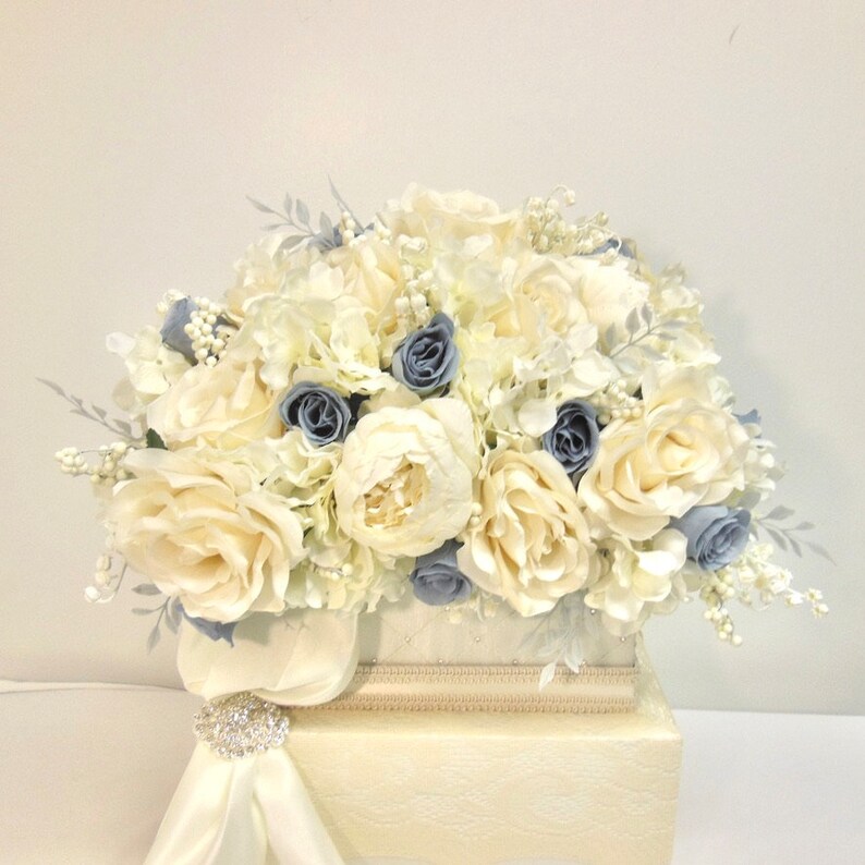 Pu&ograve; includere: Un arrangiamento floreale bianco e blu in una scatola bianca. L'arrangiamento comprende rose bianche, peonie bianche e ortensie blu.