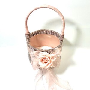 May include: A decorative pink basket with a handle, embellished with rhinestones. A pink bow and a fabric rose adorn the front. The interior is lined with pink fabric, creating an elegant and feminine design.