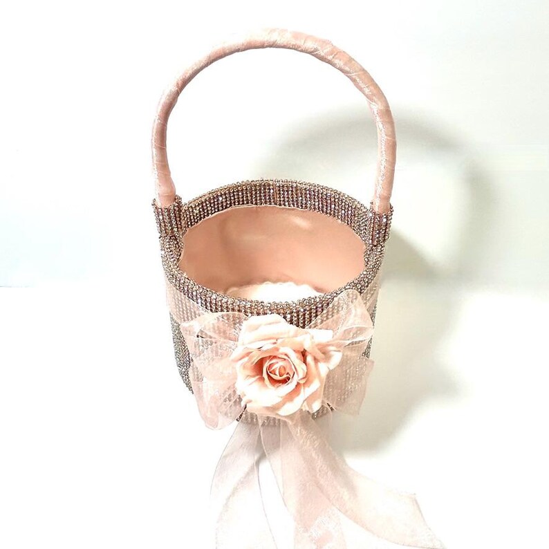 May include: A decorative pink basket with a handle, embellished with rhinestones. A pink bow and a fabric rose adorn the front. The interior is lined with pink fabric, creating an elegant and feminine design.