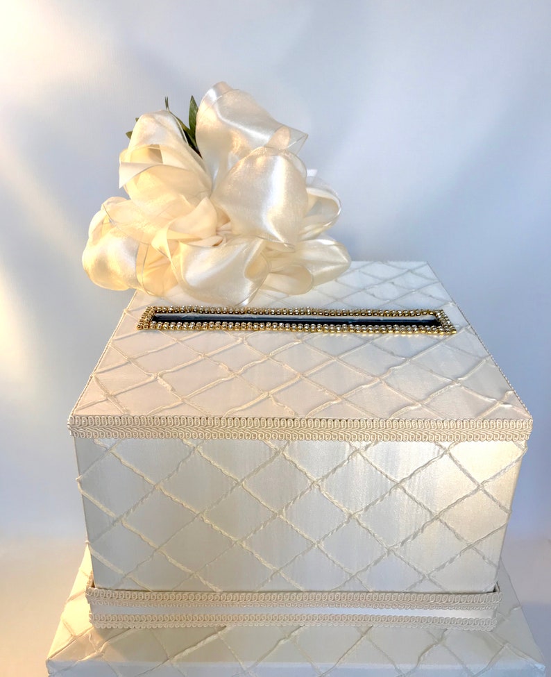 May include: White wedding card box with a large white satin bow on top. The box has a diamond pattern and a gold trim around the top.