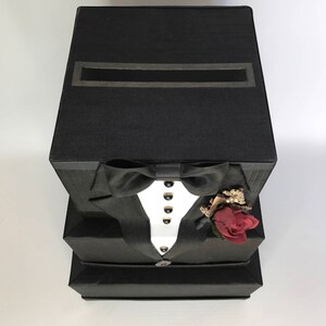 May include: A black box shaped like a tuxedo with a slot on the top for cards. The tuxedo has a white shirt, a black bow tie, and a red rose boutonniere.