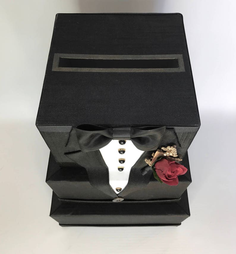May include: A black box shaped like a tuxedo with a slot on the top for cards. The tuxedo has a white shirt, a black bow tie, and a red rose boutonniere.