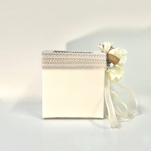 May include: A white square box with a white lace trim and a white flower with a gold center. The box is tied with a white ribbon.