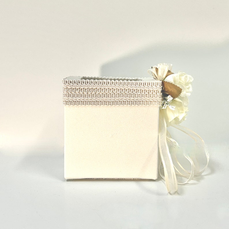 May include: A white square box with a white lace trim and a white flower with a gold center. The box is tied with a white ribbon.