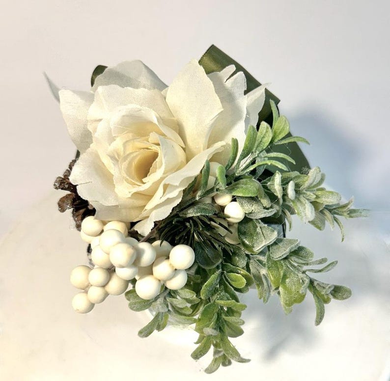 K&ouml;nnte beinhalten: Eine wei&szlig;e Kunstrose mit gr&uuml;nem Laub und wei&szlig;en Beeren in einem kleinen wei&szlig;en Topf. Die Rose ist aus Papier und hat einen zarten, vintage-Look.