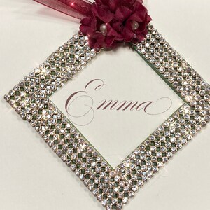 May include: A white card with a diamond-shaped frame made of clear rhinestones. The frame is decorated with a green border and a red flower with a pearl center. The name "Emma" is written in cursive inside the frame.
