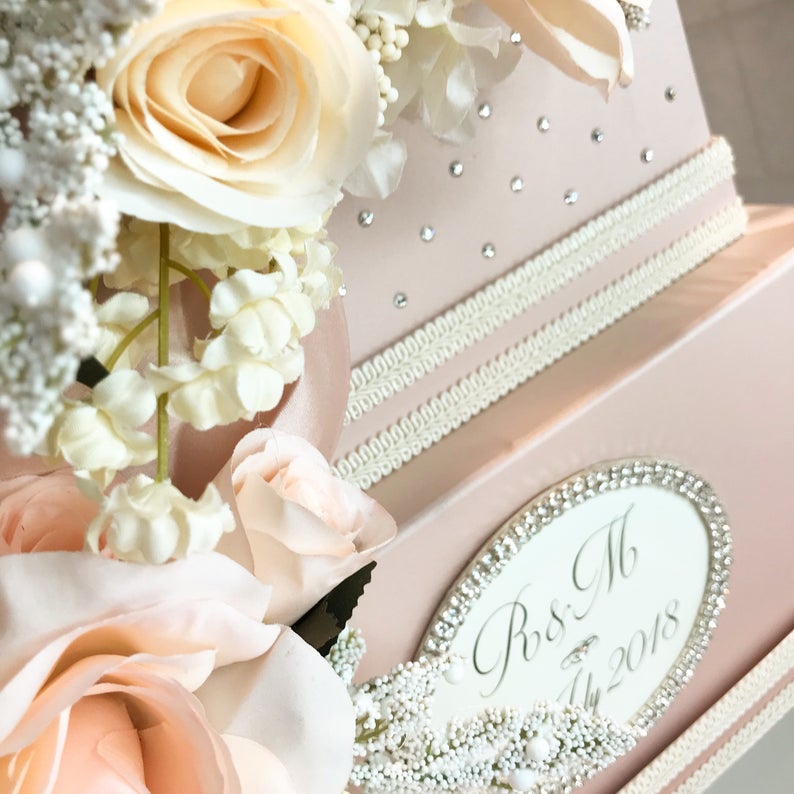 May include: A pink wedding card box with a white satin ribbon and a white oval label with the text "R&M July 2018". The box is decorated with small rhinestones and surrounded by white and peach flowers.