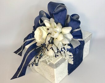 May include: A white gift box with a navy blue satin ribbon and bow. The box is decorated with white flowers and silver glitter.