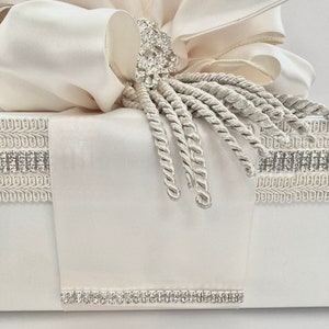 May include: Ivory-colored gift box with a large bow and rhinestone embellishments. The bow is made of layered fabric, and the box is decorated with rhinestone trim and a decorative tassel.