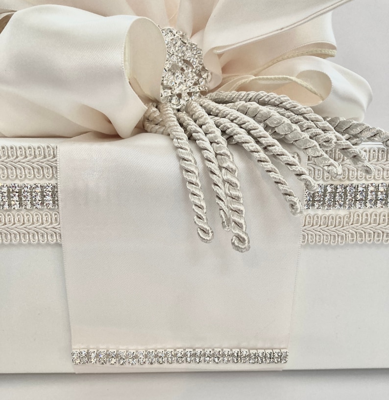 May include: Ivory-colored gift box with a large bow and rhinestone embellishments. The bow is made of layered fabric, and the box is decorated with rhinestone trim and a decorative tassel.