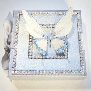 May include: A silver glitter box with a white butterfly on the front. The box is decorated with a silver rhinestone border.
