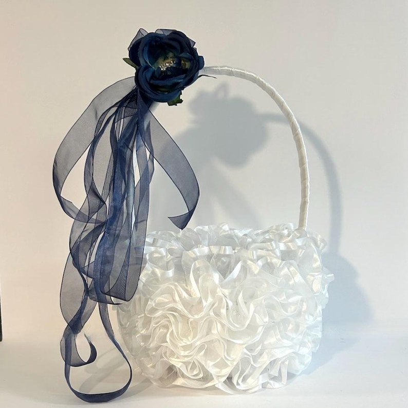 May include: A white flower girl basket with a ruffled design. The basket has a white handle and is decorated with a navy blue flower and sheer ribbons.