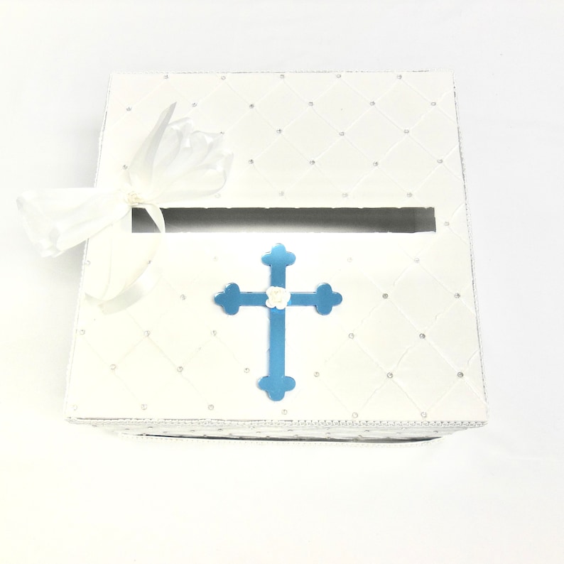 May include: White gift box with a blue cross and a white flower on top. The box has a diamond pattern and a white ribbon.
