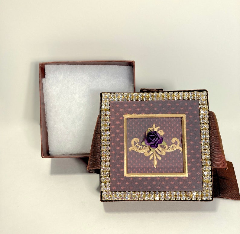 以下が含まれることがあります： 白い内装と装飾的な蓋が付いた茶色の箱。蓋は金色のデザインと紫色の花で飾られています。箱はラインストーンで縁取られています。