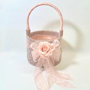 May include: A pink flower girl basket with a handle. The basket is covered in rhinestones and has a pink ribbon and a pink rose attached to the front. The basket is designed for a wedding or special event.