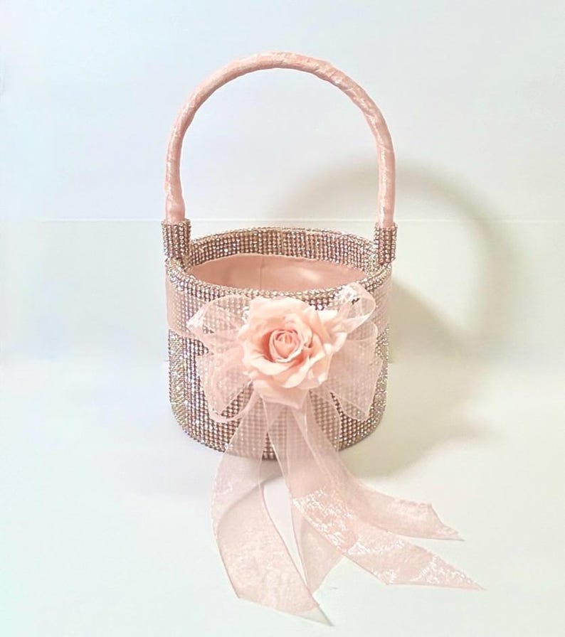 May include: A pink flower girl basket with a handle. The basket is covered in rhinestones and has a pink ribbon and a pink rose attached to the front. The basket is designed for a wedding or special event.