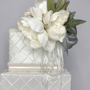 May include: Two-tiered white gift box with a quilted pattern and a white satin ribbon. The top tier has a white flower arrangement with green leaves and a silver ribbon.