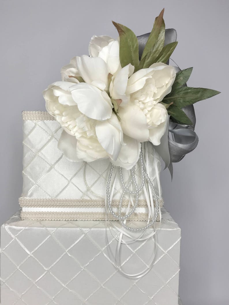May include: Two-tiered white gift box with a quilted pattern and a white satin ribbon. The top tier has a white flower arrangement with green leaves and a silver ribbon.