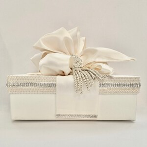 May include: Ivory-colored gift box with a large satin bow and a decorative rhinestone and tassel accent. The box is adorned with rhinestone trim and a satin ribbon. A luxurious presentation for special occasions.
