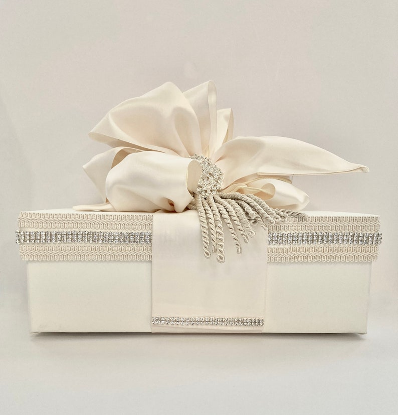 May include: Ivory-colored gift box with a large satin bow and a decorative rhinestone and tassel accent. The box is adorned with rhinestone trim and a satin ribbon. A luxurious presentation for special occasions.
