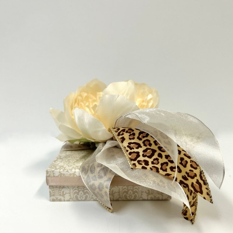 Puede incluir: Una caja de regalo blanca con un dise&ntilde;o estampado beige y blanco. La caja est&aacute; decorada con una flor de seda blanca y una cinta de estampado de leopardo marr&oacute;n y negro.