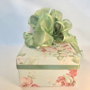 Puede incluir: Una caja de regalo floral verde con una cinta de sat&eacute;n verde y un lazo. La caja est&aacute; decorada con un patr&oacute;n floral y tiene un fondo azul claro.