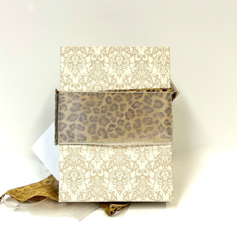 Puede incluir: Una caja blanca con un patr&oacute;n de damasco y una cinta de estampado de leopardo. La caja est&aacute; atada con un lazo.
