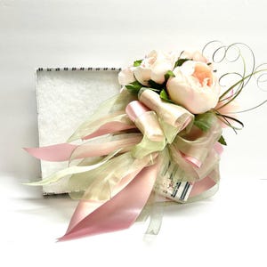 K&ouml;nnte beinhalten: Ein Blumenarrangement mit hellrosa Pfingstrosen, gr&uuml;nen Bl&auml;ttern und transparenten B&auml;ndern in Rosa und Gr&uuml;n. Das Arrangement ist mit einer gro&szlig;en Schleife gebunden und befindet sich neben einer wei&szlig;en Box.