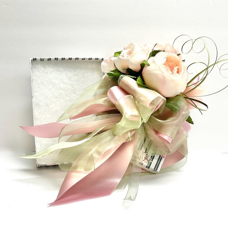 K&ouml;nnte beinhalten: Ein Blumenarrangement mit hellrosa Pfingstrosen, gr&uuml;nen Bl&auml;ttern und transparenten B&auml;ndern in Rosa und Gr&uuml;n. Das Arrangement ist mit einer gro&szlig;en Schleife gebunden und befindet sich neben einer wei&szlig;en Box.