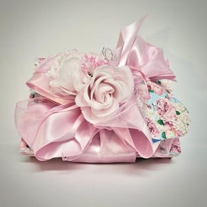 May include: A rectangular box decorated with a light blue floral pattern featuring pink roses. The top is adorned with a large, voluminous light pink satin ribbon bow and several artificial pale pink rose blossoms.