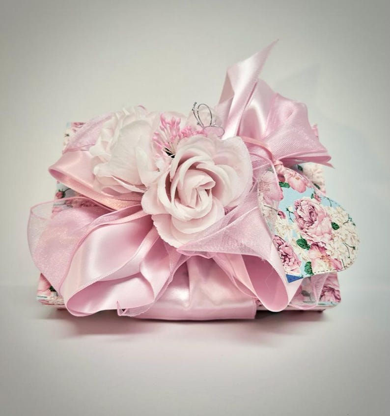 May include: A rectangular box decorated with a light blue floral pattern featuring pink roses. The top is adorned with a large, voluminous light pink satin ribbon bow and several artificial pale pink rose blossoms.