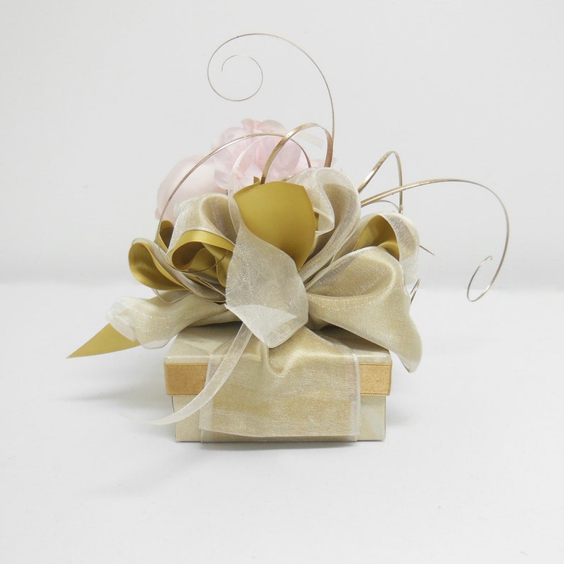 K&ouml;nnte beinhalten: Eine kleine Geschenkbox, die in beigefarbenes Papier eingewickelt ist, mit einem goldenen Band und einer rosa Blume oben drauf.