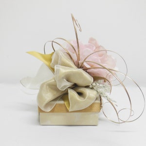 K&ouml;nnte beinhalten: Eine kleine, beige Geschenkbox mit einem goldenen Band und einem gro&szlig;en, rosafarbenen Blumenarrangement obenauf. Das Blumenarrangement besteht aus Seidenblumen und hat einen goldenen Draht-Stiel.