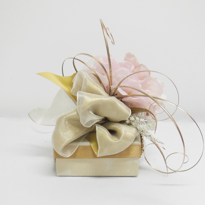 K&ouml;nnte beinhalten: Eine kleine, beige Geschenkbox mit einem goldenen Band und einem gro&szlig;en, rosafarbenen Blumenarrangement obenauf. Das Blumenarrangement besteht aus Seidenblumen und hat einen goldenen Draht-Stiel.