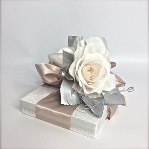 May include: A white gift box with a beige satin ribbon and a single white rose with silver leaves.
