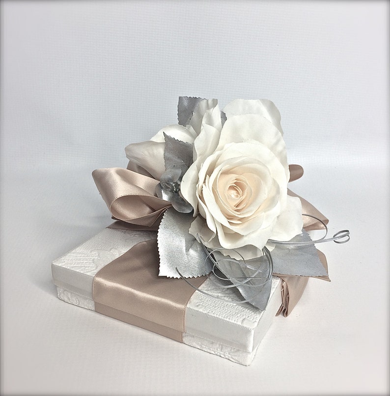 May include: A white gift box with a beige satin ribbon and a single white rose with silver leaves.